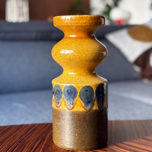 Może przedstawiać: Wazon ceramiczny w stylu vintage o pękatym kształcie i teksturowanej powierzchni. Wazon charakteryzuje się gradientem kolorów, przechodzącym od ciemnobrązowej podstawy do żółto-pomarańczowej góry. Niebieskie wzory w kształcie łez zdobią środkową część.