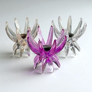 May include: Three clear and purple lotus flower-shaped candle holders. The candle holders have a clear, petal-like design with a dark center. The central candle holder is a vibrant purple color, while the other two are clear.