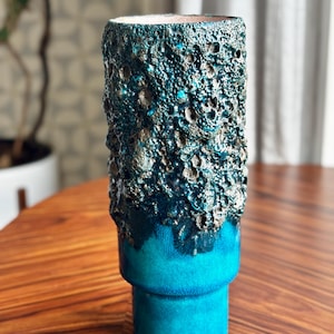 Könnte beinhalten: Eine hohe, zylindrische Keramikvase mit einer strukturierten, dunkelblauen Außenseite und einem glatten, hellblauen Sockel. Der obere Teil hat eine raue, zerklüftete Oberfläche, während der Sockel eine einfarbige, glatte Farbe hat. Die Vase steht auf einem Holztisch.