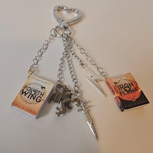 Puede incluir: Un llavero plateado con cierre en forma de corazón, con dijes de libros en miniatura, un dragón, una espada y un rayo. Los libros se titulan "Fourth Wing" e "Iron Flame" de Rebecca Yarros. El dije de dragón es de color bronce.