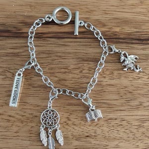 Può includere: Bracciale con ciondoli color argento con chiusura a toggle. Il bracciale presenta diversi ciondoli, tra cui un acchiappasogni, un libro aperto, un drago e un ciondolo rettangolare con la scritta "I LOVE BOOKS".