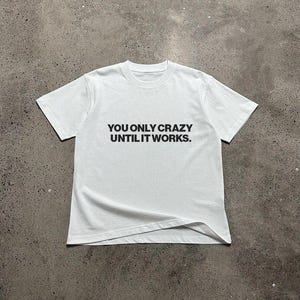Könnte beinhalten: Weißes T-Shirt mit schwarzem Text, der "YOU ONLY CRAZY UNTIL IT WORKS." lautet.