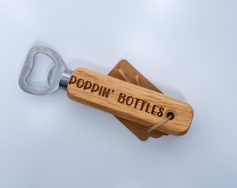 May include: A bottle opener with a light brown wooden handle featuring the engraved text POPPIN' BOTTLES. The metal opener head is silver-colored, and a small brown rectangular tag is attached to the handle with a thin piece of twine.