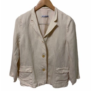 Może przedstawiać: Kremowy blazer z lnu z metką Jil Sander. Blazer ma jedno guzowe zapięcie i dwie naszywane kieszenie.