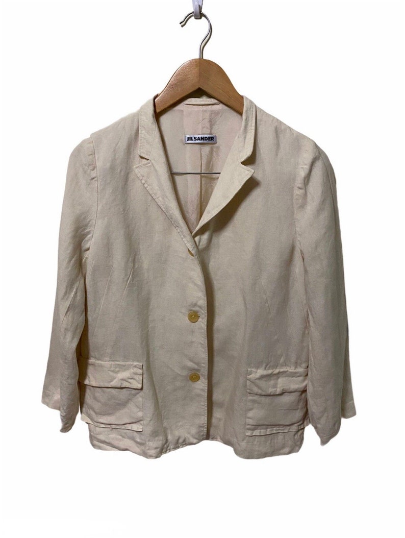 Może przedstawiać: Kremowy blazer z lnu z metką Jil Sander. Blazer ma jedno guzowe zapięcie i dwie naszywane kieszenie.