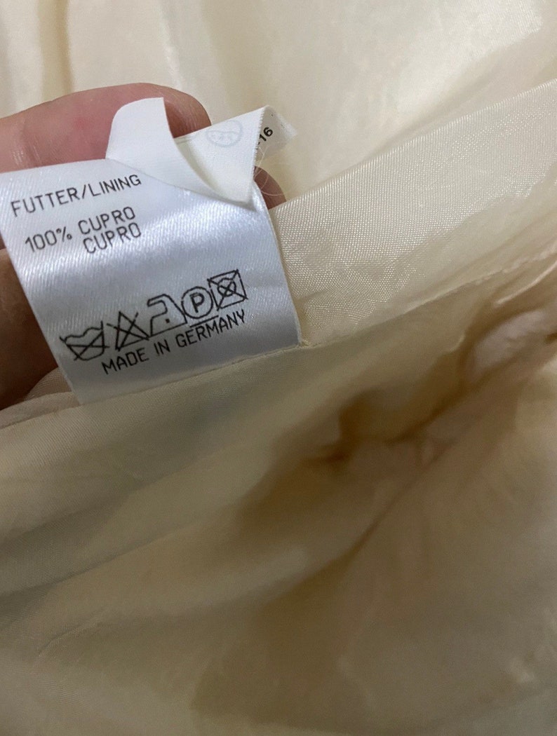 Może przedstawiać: Biała metka odzieżowa z tekstem "FUTTER/LINING 100% CUPRO CUPRO" i "MADE IN GERMANY". Metka zawiera r&oacute;wnież etykietę pielęgnacyjną z symbolami prania, prasowania i czyszczenia chemicznego.
