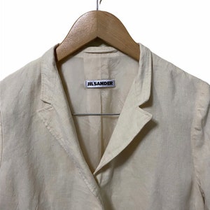 Może przedstawiać: Kremowy blazer z lnu z metką Jil Sander na kołnierzyku.