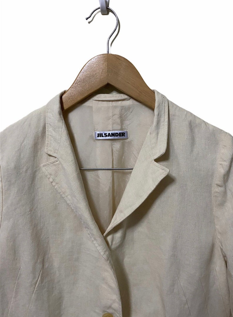 Może przedstawiać: Kremowy blazer z lnu z metką Jil Sander na kołnierzyku.