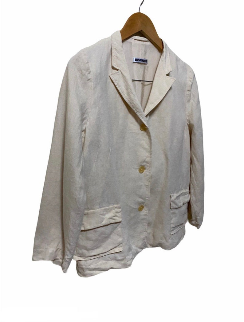 Może przedstawiać: Kremowy blazer z lnu z metką Jil Sander. Blazer ma kołnierz szalowy, trzy guziki i dwie naszywane kieszenie.