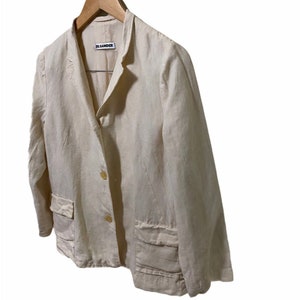 Może przedstawiać: Kremowy blazer z lnu z pojedynczym zapięciem na guzik i dwiema naszywanymi kieszeniami. Blazer ma metkę Jil Sander.