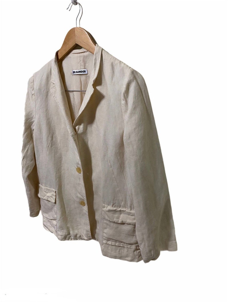 Może przedstawiać: Kremowy blazer z lnu z pojedynczym zapięciem na guzik i dwiema naszywanymi kieszeniami. Blazer ma metkę Jil Sander.