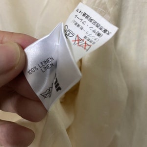 Może przedstawiać: Biała metka z tkaniny z czarnym tekstem, kt&oacute;ry brzmi "100% LEINEN LINEN". Druga biała metka z czarnym tekstem jest częściowo widoczna. Tekst brzmi "自然乾燥 中 (タンブラー乾燥禁止) (株)サン・フレール 大阪市西区阿波座1-4-16".