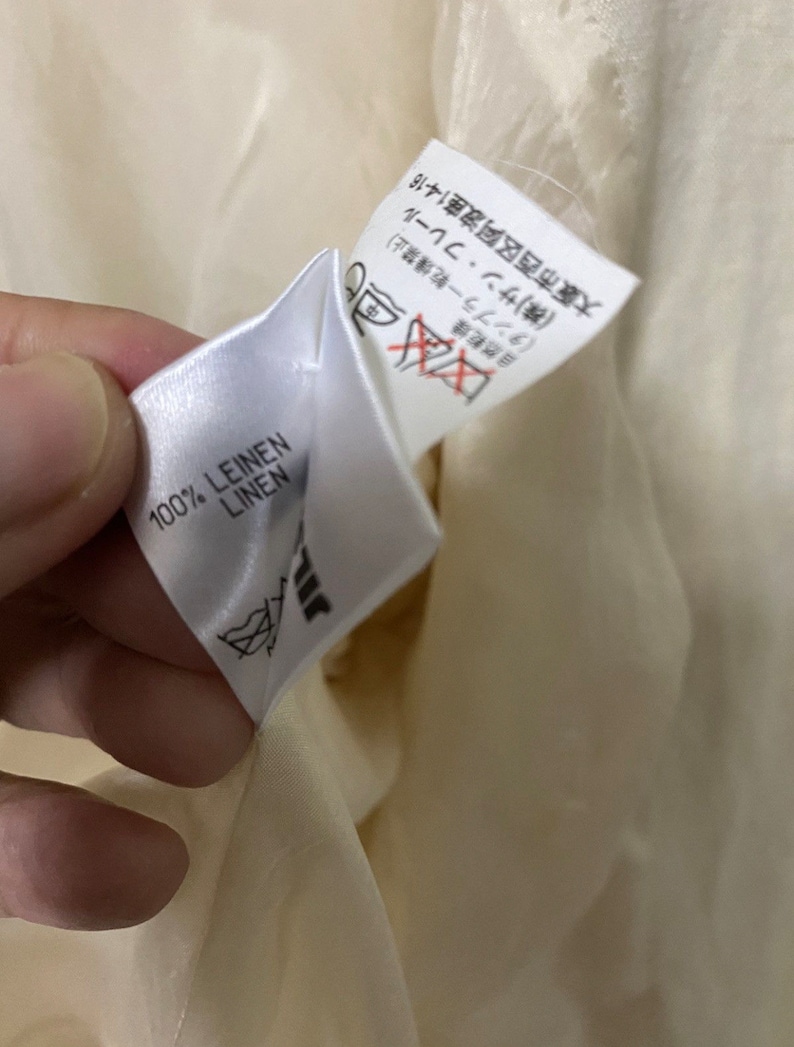 Może przedstawiać: Biała metka z tkaniny z czarnym tekstem, kt&oacute;ry brzmi "100% LEINEN LINEN". Druga biała metka z czarnym tekstem jest częściowo widoczna. Tekst brzmi "自然乾燥 中 (タンブラー乾燥禁止) (株)サン・フレール 大阪市西区阿波座1-4-16".