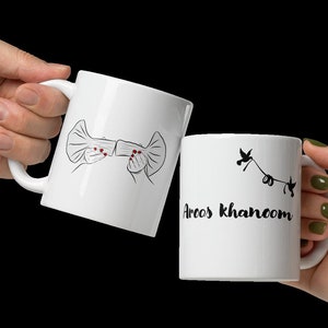 May include: Two white ceramic mugs with black designs. One mug has a drawing of two hands holding a bow. The other mug has a drawing of a bird with the text "Areos Khanom".