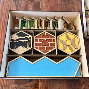 May include: Wooden board game organizer with compartments for cards and game pieces. The organizer features hexagonal tiles with desert, brick, and wheat designs. A blue, angular piece is at the bottom. The cards are in the top section.
