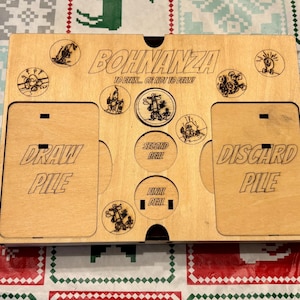 Könnte beinhalten: Karten-Organizer für das Bohnanza-Kartenspiel aus Holz. Der Organizer hat Fächer für den Ziehstapel und den Ablagestapel. Der Text "Bohnanza" und "To Deal... Or Not To Deal!" sind sichtbar. Der Organizer hat Cartoon-Illustrationen.