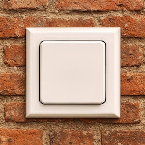 Op de afbeelding: Een witte lichtschakelaar met een vierkant ontwerp, geplaatst in een wit frame, gemonteerd op een rode bakstenen muur. De schakelaar heeft een zwarte rand en een gestikt detail op het frame.