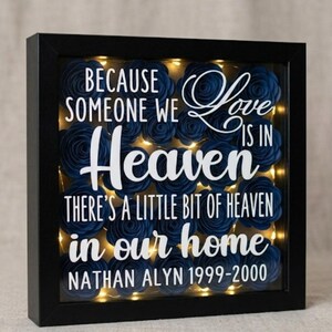 May include: A square black shadow box frame containing dark blue paper roses and warm LED string lights. The glass front features white text reading: Because someone we love is in heaven there's a little bit of heaven in our home Nathan Alyn 1999-2000.
