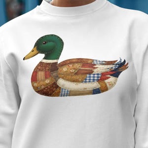 Peut inclure: Sweat-shirt blanc orné d'une application de canard. Le canard a une tête verte, un bec jaune et un corps fait de patchs de tissu marron, bleu et rouge. Le design donne un aspect matelassé. Vêtement confortable et décontracté.