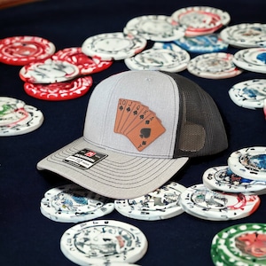 May include: A gray and black trucker hat with a leather patch featuring a royal flush of spades. The hat is lying on a blue felt surface with poker chips scattered around it.
