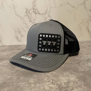 May include: A gray and black trucker hat with a black patch featuring the number 777 in a silver font. The patch has a black border and a ring of silver dots around the outside.