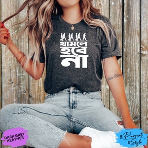 May include: A dark grey heather t-shirt with a white graphic design featuring a line of stick figures walking with the text "Mama I'm Not" in Bengali.