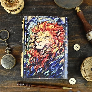 May include: A leather-bound journal with a stained glass design of a lion's head. The lion's mane is red, orange, and yellow, and the background is blue and purple.