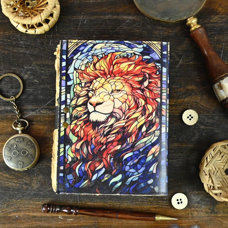 May include: A leather-bound journal with a stained glass design of a lion's head. The lion's mane is red, orange, and yellow, and the background is blue and purple.
