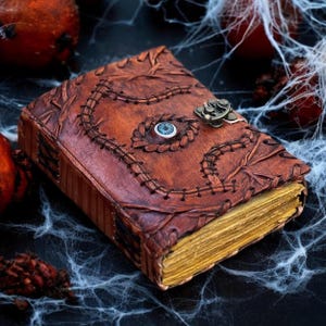 Op de afbeelding: Een bruin leren boek met een gestikt ontwerp en een blauw oog in het midden. Het boek heeft een metalen sluiting en vergeelde pagina's. De omslag is in reliëf met ingewikkelde patronen, wat een vintage of fantasiethema oproept.