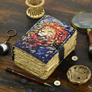 May include: A leather-bound journal with a stained glass design featuring a lion's head. The journal has a clasp closure and a leather strap.
