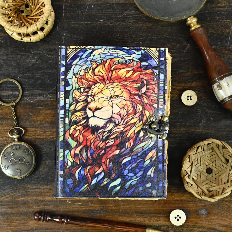 May include: A leather-bound journal with a colorful stained glass design of a lion's head. The journal has a metal clasp closure.
