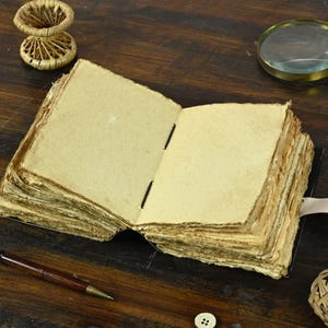 May include: An open, blank journal with aged, yellowed pages. The journal has a leather cover and a leather strap closure. The journal is lying open on a wooden table with a magnifying glass, a pocket watch, and a pen nearby.
