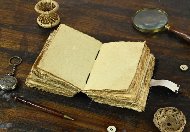 May include: An open, blank journal with aged, yellowed pages. The journal has a leather cover and a leather strap closure. The journal is lying open on a wooden table with a magnifying glass, a pocket watch, and a pen nearby.