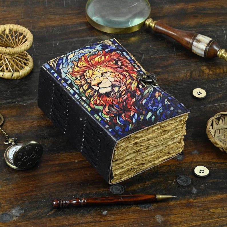 May include: A black leather journal with a stained glass lion design on the cover. The journal has a metal clasp and is closed. The journal is sitting on a wooden surface with other objects around it.