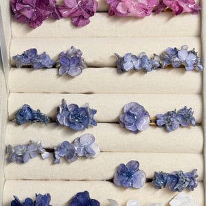 Può includere: Una collezione di anelli floreali in un portagioie. Gli anelli presentano vari disegni floreali viola, blu e bianchi. La scatola è beige e gli anelli sono disposti in file. I fiori sembrano essere fatti di resina.