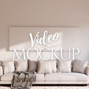 以下が含まれることがあります： リビングルームに置かれた白いソファ。クッションと毛布が置かれている。ソファの上の壁には、白いフレームの中に「Video Mockup」という文字が書かれたものが飾られている。