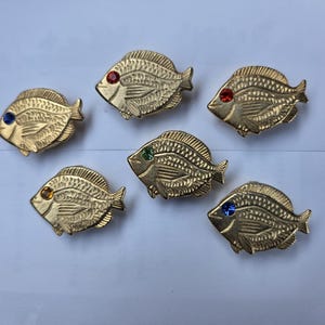 Peut inclure: Six broches en métal doré en forme de poisson, chacune présentant un motif d'écailles texturé et un petit strass serti en guise d'œil. Les strass sont de différentes couleurs, notamment rouge, bleu, vert et ambre, disposés en formation éparse.