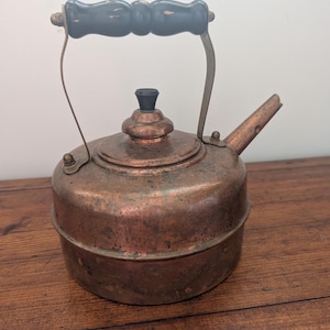 May include: A vintage copper kettle with a black wooden handle and spout. The kettle has a round body and a lid with a black knob. The handle is attached to the kettle with metal brackets. The kettle is sitting on a wooden surface.