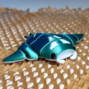 Può includere: Una statuetta di manta turchese e bianca con un occhio nero. La statuetta è stampata in 3D e presenta un design dettagliato. La manta si trova su una superficie marrone a nido d'ape.