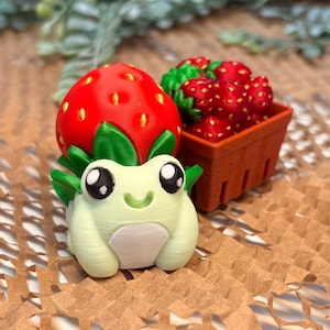 May include: A small, light green, 3D-printed frog figurine with a large red strawberry on its head, featuring yellow seeds and green leaves. Beside the frog is a miniature brown basket filled with several small, detailed red strawberries.