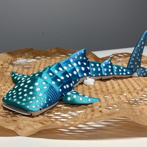 Puede incluir: Un modelo detallado y articulado de tiburón ballena en tonos turquesa y azul, con lunares blancos. El tiburón de juguete está sobre papel marrón. El modelo tiene un cuerpo segmentado y aletas.