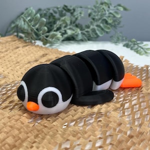 Puede incluir: Un juguete de pingüino articulado impreso en 3D en negro, blanco y naranja. El pingüino tiene un cuerpo segmentado, lo que permite una postura flexible. El juguete está sobre una superficie marrón texturizada, con follaje verde en el fondo.