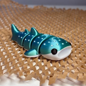 May include: A teal and white articulated whale shark toy with white spots. The toy has black eyes and a white mouth. The toy is resting on a brown, honeycomb-patterned surface.