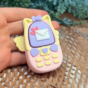 May include: A pastel pink and yellow mobile phone accessory featuring a lavender screen with a white envelope icon, a small lavender bow at the top, and yellow heart-shaped buttons. The device has small yellow wings on the sides.