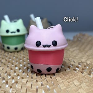 May include: A pink and red boba tea cup-shaped keyring with a cat face design. The keyring has a white straw and black boba pearls. The text "Click!" is in the background.