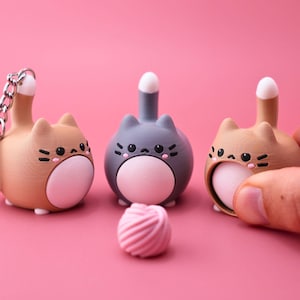 May include: Three small, rounded cat-shaped figurines with upright tails and white bellies. Two are light brown and one is gray. Each has black eyes, whiskers, and pink cheek spots. One brown cat features a silver metal keychain ring attached.