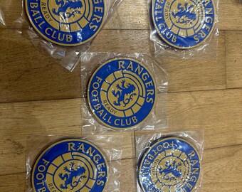 Puede incluir: Cinco insignias del Rangers Football Club en embalaje transparente. Cada insignia circular presenta el logotipo del equipo, un león dorado y las palabras "Rangers" y "Football Club". Las insignias son azules y doradas.