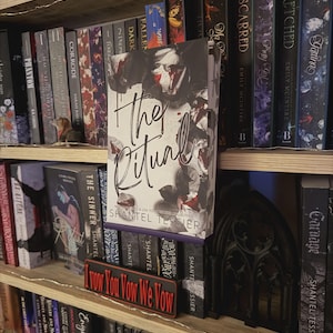 Op de afbeelding: Een boek getiteld The Ritual van Shantel Tessier met een zwart-witte bloemenomslag, tentoongesteld op een houten boekenplank tussen andere boeken. Een klein rechthoekig bordje met de tekst I Vow You Vow We Vow staat voor het boek.