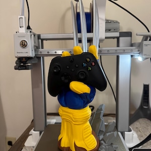 May include: A 3D printed controller stand shaped like a yellow and blue gloved hand with three long silver metallic claws extending upwards. The hand is gripping a black wireless game controller, and a grey rocky base is attached to the bottom.