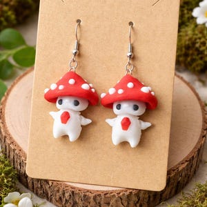 May include: A pair of whimsical mushroom-shaped earrings. Each earring features a red cap with white spots atop a white body with outstretched arms. The earrings are displayed on a brown card, set against a backdrop of moss and flowers.